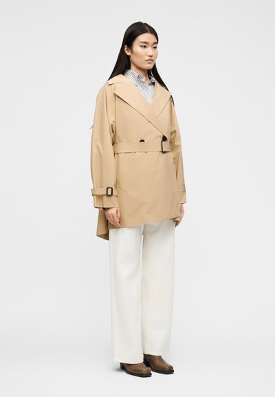 Γυναίκα με μακριά σκούρα μαλλιά που φοράει ένα μπεζ μπουφάν με ζώνη, ριγέ πουκάμισο, λευκό παντελόνι φαρδιάς γραμμής και καφέ μπότες, στέκεται μπροστά σε απλό φόντ ο.