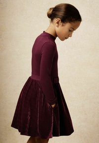 Bordová šaty s priliehavým dlhým rukávom a rozšírenou zamatovou sukňou, doplnené o zodpovedajúci pás a bočné vrecká.