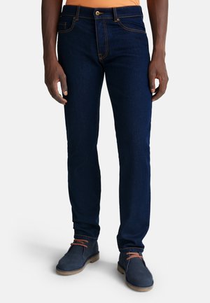 Jeans de mezclilla azul oscuro con corte ajustado, con costuras naranjas y diseño estándar de cinco bolsillos. Llevados con botas de tobillo grises.