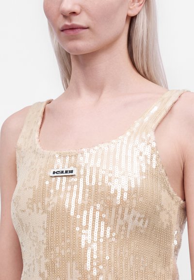 Gros plan d'une personne portant un débardeur beige à sequins avec un col carré et une petite étiquette rectangulaire de la marque Ksubi sur la poitrine.