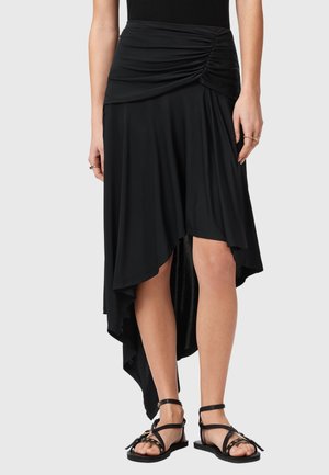 Schwarzer asymmetrischer Rock mit geraffter Detail am Bund, getragen mit schwarzen Riemchensandalen an einer stehenden Frau.