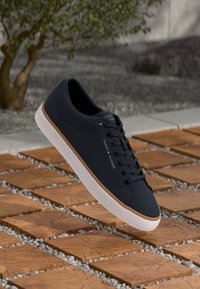 Sort Tommy Hilfiger sneakers med hvid sål og brune detaljer på trækvadrater med små sten imellem dem udendørs.