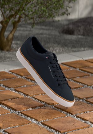 Crni Tommy Hilfiger tenisice s bijelim đonom i smešnim detaljima na drvenim kvadratnim pločama sa malim kamenčićima između njih na otvorenom.