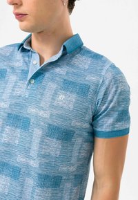 Blå mønstrede polo-shirt med krave, korte ærmer og subtil tekstur. Har et lille logo på brystet.