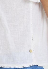 Haut en lin blanc avec un tissu texturé, présentant un bouton décoratif et un ourlet festonné. Le design comprend des coutures délicates le long de la couture.