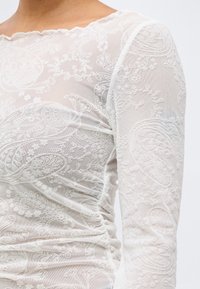 Γυναίκα που φοράει λευκό διάφανο δαντελένιο τοπ με λουλουδάτα και παϊέτα μοτίβα, με μακριά μανίκια και σούρες στις πλευρές.