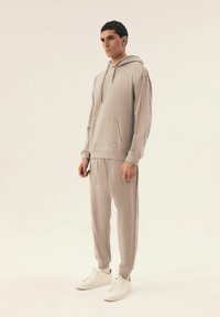 Jasnoszara bluza z kapturem i dopasowane dresy wykonane z miękkiego materiału. Posiada kieszeń z przodu, regulowany kaptur na sznurki oraz ściągacze przy mankietach.