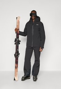 Schwarze Skijacke und -hose mit Kapuze, hält beige und schwarze Ski mit Bindungen. Große schwarze Skibrillen bedecken die Augen.