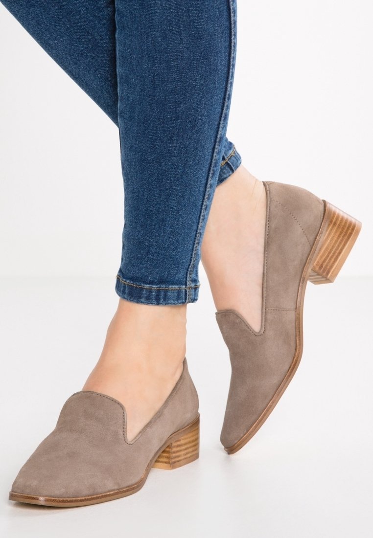 Chaussures slip-on en suède gris avec un bout arrondi et un design en V. Le talon en bois effilé ajoute un contraste. Portées avec un jean bleu ajusté.