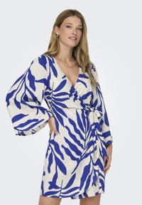 Wikkeljurk met een blauw bladpatroon op een witte achtergrond; de stoffen hebben een gladde textuur, met klokmouwen en een strikdetail in de taille.