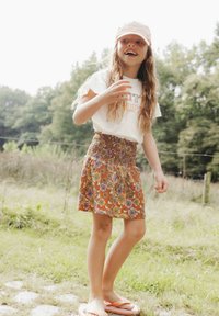 Meisje in een witte T-shirt en een oranjerode bloemenrok met een patroon. Ze heeft lang haar en staat op gras, met roze slippers aan.