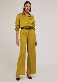 Satin, långärmad skjorta och vida byxor i klar senapsgul färg; bälte i brunt läder; modellen bär mörkröda högklackade skor.