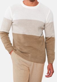 Pull en maille avec un design à blocs de couleurs en blanc, beige clair et brun clair. Texture structurée avec poignets et ourlet côtelés. Le mannequin porte un pantalon beige.
