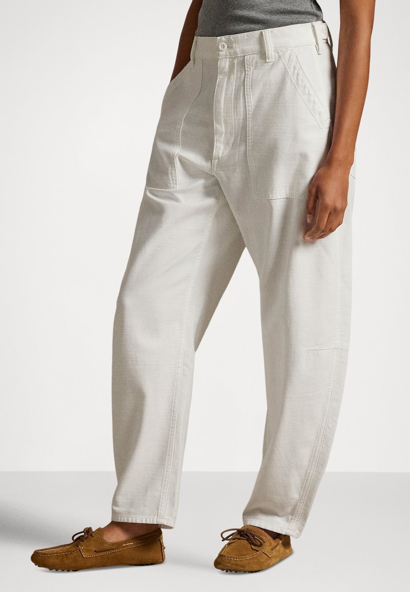 Person iført off-white bukser med avslappet passform og frontlommer, og brune mokasinsko i semsket skinn, stående mot en ensfarget bakgrunn.