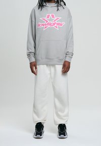 Sudadera gris con logo de estrella rosa y bolsillo frontal, combinada con pantalones de chándal blancos y zapatillas deportivas negras con detalles blancos.