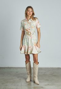 Blumenkleid aus leichtem Stoff, mit kurzen Ärmeln, tailliertem Schnitt und ausgestelltem Saum. Kombiniert mit kniehohen beigen Cowboystiefeln.