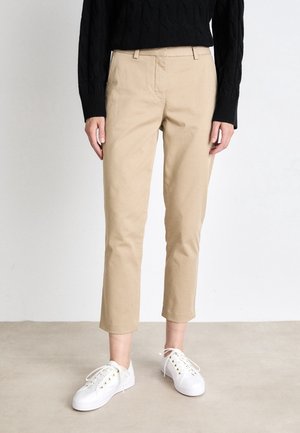 Osoba ubrana w beżowe krótkie spodnie, białe sportowe buty i czarny sweter z długim rękawem, stoi na jasnej podłodze przy bladej ścianie.