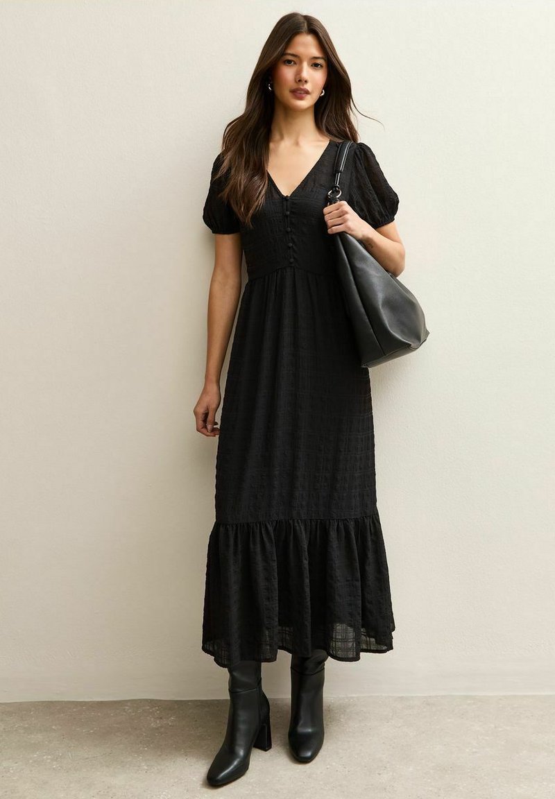 Woman wearing a black midi dress with puff sleeves and buttons, black heeled boots, carrying a large black shoulder bag against a plain wall.