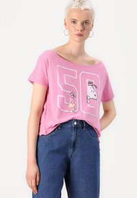 Růžové bavlněné tričko s designem přes rameno, které zobrazuje grafiku Snoopyho a květiny, s číslem "50" v bílé kontuře. Nosené s modrými džíny.