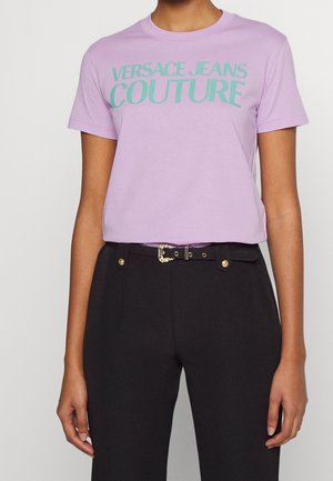 Osoba nosící levandulové tričko Versace Jeans Couture zastrčené do černých kalhot s dekorativním páskem se zlatou sponou, viditelná od krku po boky.