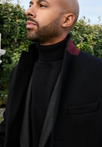Abrigo negro con un cuello alto, llevado sobre un suéter negro de cuello alto. El abrigo presenta un acento burdeos cerca del hombro. Textura de tela suave.