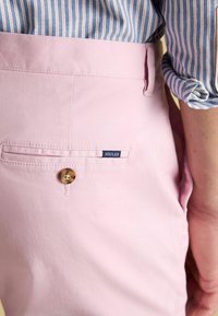 Lichtroze broek met een achterzak met knoopsluiting en een klein blauw label met de tekst "JOULES." De stof heeft een gladde textuur.