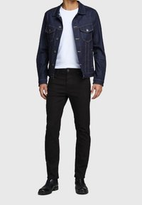 Chaqueta de mezclilla oscura con botones plateados, llevada sobre una camiseta blanca, combinada con pantalones negros ajustados y botas negras al tobillo.