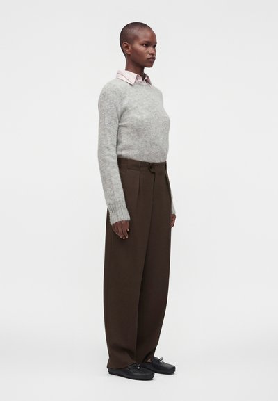Personne de profil portant un pull gris sur une chemise rose, un pantalon large marron et des chaussures noires, sur un fond blanc uni.