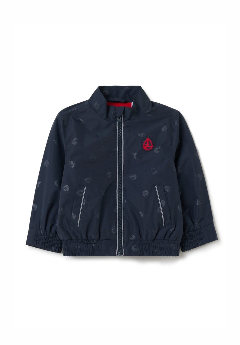 Chaqueta cortavientos en color azul marino con cuello alto, cierre de cremallera, puños elásticos y un sutil patrón de íconos de veleros. Presenta un detalle del logotipo en rojo.