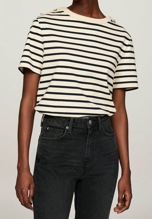 Person iført en creme- og sortstribet kortærmet top med guldknapper på skuldrene og sort højtaljet jeans.