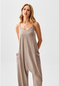 Brauner ärmelloser Jumpsuit mit lockerem Schnitt, dünnen Trägern und zwei Taschen vorne, aus weichem, strukturiertem Stoff.