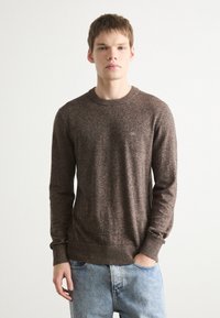 Brązowy sweter z dzianiny, z okrągłym dekoltem, o fakturowanej tkaninie i ściągaczach. Posiada małe logo na piersi, zestawiony z jasnoniebieskimi dżinsami.