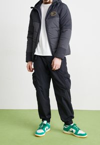 Svart puffjacka med huva och guldlogotyp, ihopparad med svarta cargo-byxor. Skorna inkluderar gröna och vita sneakers med en gummisula.