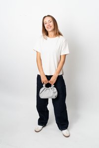 Ung kvinna som ler, klädd i vit t-shirt, mörka, lösa jeans och vita sneakers, håller en liten grå handväska med båda händerna framför sig.