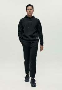 Muž v čiernej mikine s kapucňou, zodpovedajúcich jogger nohaviciach a čiernych teniskách, kráča smerom k fotoaparátu proti jednoliatej svetlej pozadí.