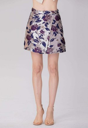 Jupe courte portefeuille fleurie avec des fleurs violettes et bleues, portée avec des talons nude à bride cheville, montrant les jambes de la taille vers le bas sur un fond uni.