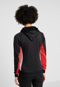 Persona con capelli ricci che indossa un hoodie nero con pannelli rossi e bianchi sulle maniche, visto da dietro su uno sfondo chiaro e semplice.