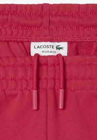 Pantalones cortos rojos de cintura elástica con cordón. Presenta la etiqueta del logo de Lacoste y un diseño de corte regular. Tejido suave con una textura blanda.
