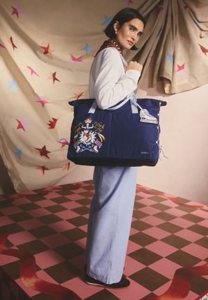 Modrá látková taška přes rameno s grafickým potiskem. Má dvojité rukojeti, stahovací šňůrku a dekorativní přívěsek ve tvaru skateboardu. Texturovaný povrch.