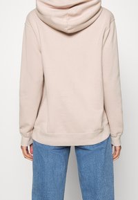 Haut à capuche beige clair avec cordon de serrage, poignets et ourlet côtelés. Associé à un jean bleu texturé. La vue de dos montre des caractéristiques de design minimales.