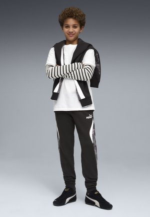 Ragazzo con capelli ricci in piedi con le braccia incrociate, indossa una maglietta bianca con maniche a righe nere e bianche, pantaloni neri Puma e scarpe da ginnastica Puma nere.