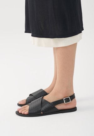 Zwarte gebreide sandalen met kruislingse banden, een platte zool en een enkelbandje met een zilveren gespdetail.