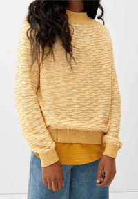 Personne portant un pull jaune texturé en maille par-dessus une chemise moutarde et un jean bleu clair, debout devant un fond clair uni.