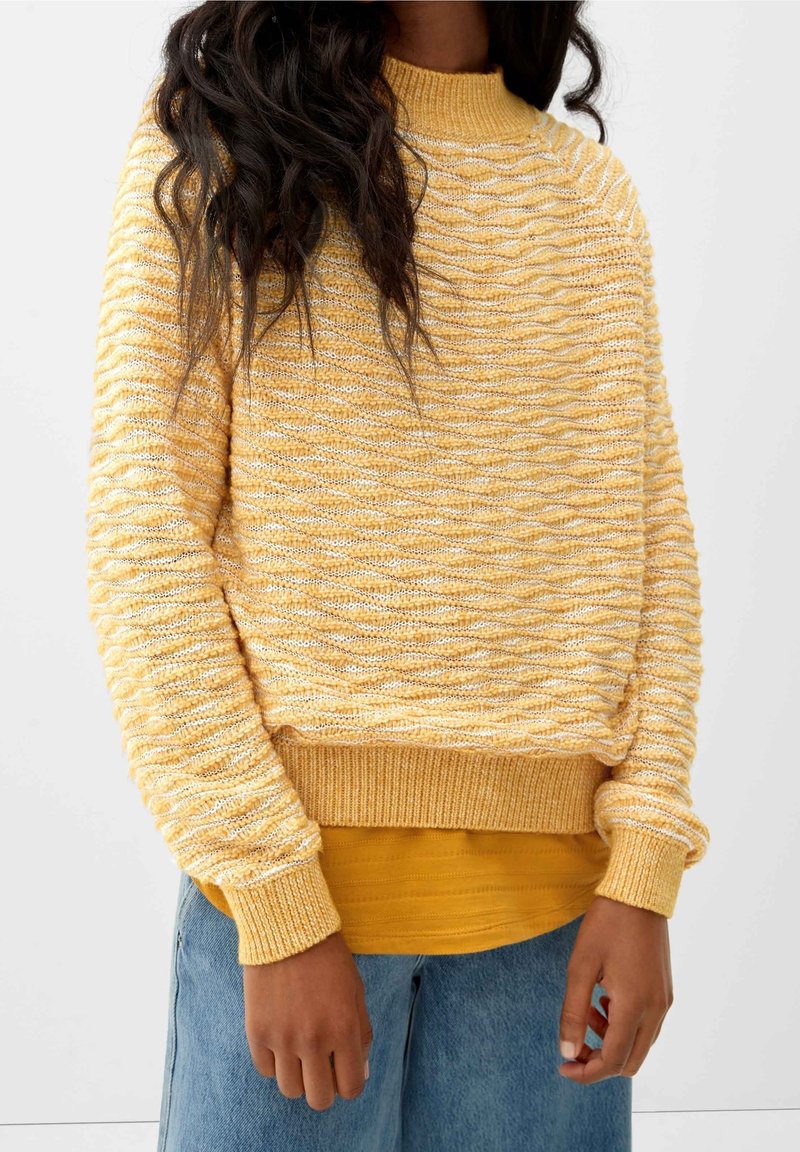 Personne portant un pull jaune texturé en maille par-dessus une chemise moutarde et un jean bleu clair, debout devant un fond clair uni.