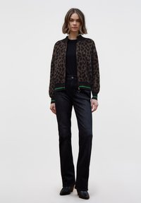 Hnědý cardigan se vzorem leopardí kůže a zeleným proužkovaným lemem, nošený přes černý top. Kombinováno s tmavými džínami a černými kotníkovými botami na podpatku.