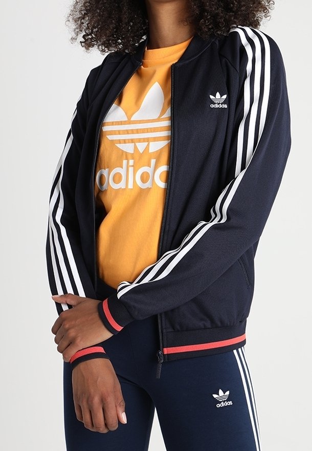 Mörkblå zip-up jacka med vita ränder på ärmarna, röda detaljer vid fållen, över en orange t-shirt med en vit Adidas-logotyp.