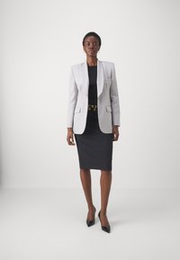 Blazer ajustado gris claro sobre un vestido negro ceñido, acentuado con un cinturón de cadena dorada. Tacones negros de punta afilada completan el look.