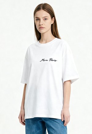 Jeune femme aux cheveux bruns et raides, portant un t-shirt blanc oversize avec l'inscription "Mira Paris" et un jean bleu, debout devant un fond uni.