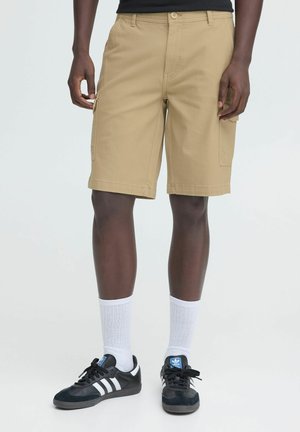 Male model wearing beige cargo shorts, white mid-calf socks, and black Adidas sneakers against plain background.