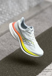 Weißer Laufschuh mit Obermaterial aus Mesh, kontrastierenden gelben und orangen Akzenten an der Sohle und einem strukturierten, leichten Design.